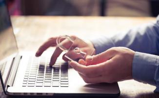 Am Laptop arbeitende Hände, eine Hand hält Glasschloss